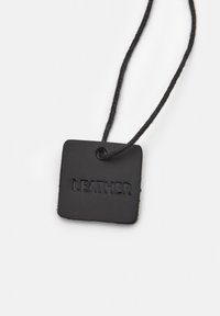 Fekete bőr címke négyzet alakban, a "LEATHER" felirattal dombornyomva. Fekete zsinórral rögzítve. Simább textúra, minimalista dizájn.
