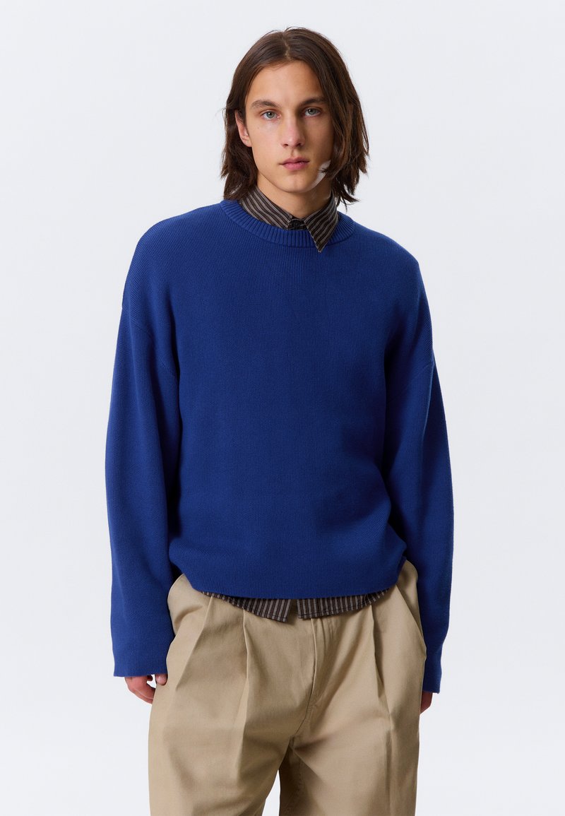 Pull en maille bleu avec un col rond, des manches longues et une coupe décontractée. Porté par-dessus une chemise à col rayée ; associé à un pantalon beige.