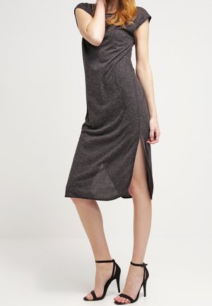 Vrouw draagt een donkergrijze midi-jurk met korte mouwen en een split tot boven de knie, gecombineerd met zwarte enkellaarzen met hoge hak en enkelriempjes.