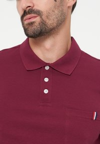 Homme avec une barbe courte portant un polo bordeaux avec trois boutons blancs et une poche poitrine avec une petite étiquette rouge, blanche et bleue.