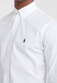 Mężczyzna ubrany w białą koszulę z guzikami, z małym haftem logo gracza polo w kolorze granatowym na piersi.