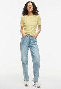 Top amarillo de manga corta acanalado, jeans de talle alto de color azul claro y tacones abiertos blancos. La modelo está de pie con las manos en los bolsillos.