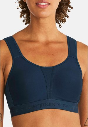 Vrouw die een donkerblauwe sportbeha draagt met dikke schouderbanden en ademende stofpanelen, waarbij haar bovenste borst en nek zichtbaar zijn.