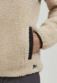 Beige fleece jacket with a textured, fluffy surface; features black trim along the cuffs and hem; includes side hand pockets.