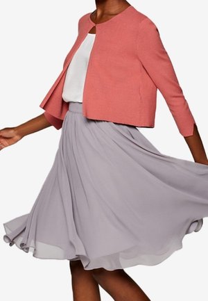 Vrouw draagt een koraalkleurig cropped jasje over een wit topje en een vloeiende lichtgrijze midi-rok, halverwege een draai waarbij de beweging van de rok zichtbaar is.