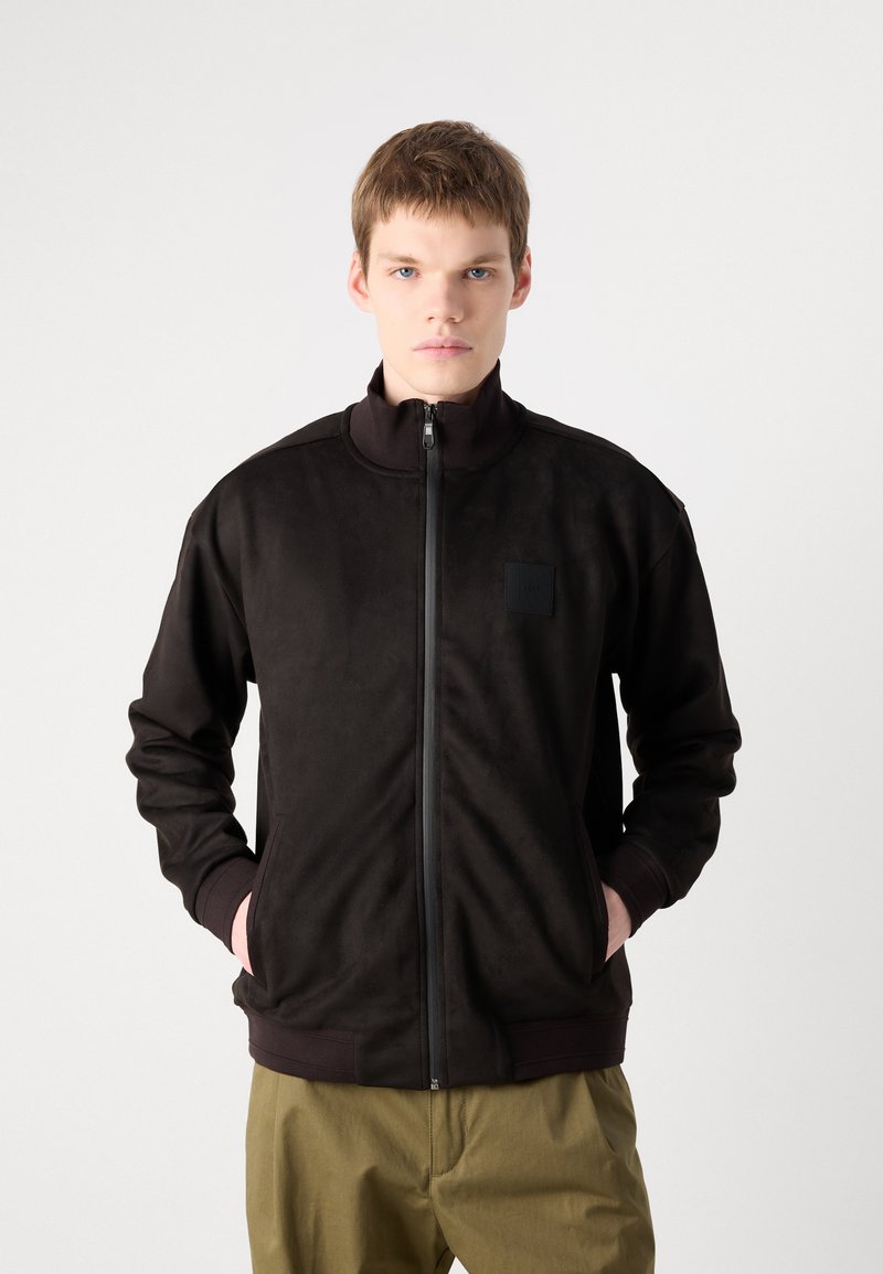 Schwarze Kapuzenjacke aus weichem Stoff, mit hohem Kragen, seitlichen Taschen und einem strukturierten quadratischen Patch auf der linken Brust.
