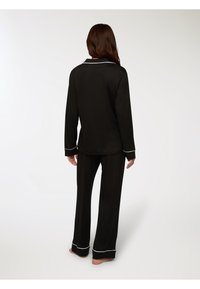 Donna in piedi a piedi nudi che indossa un pigiama lungo nero con piping bianco, vista da dietro su uno sfondo chiaro e semplice.