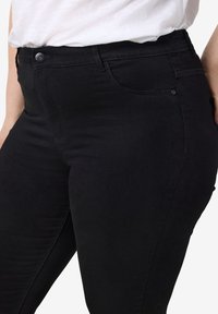 Gros plan d'une personne portant un jean noir ajusté associé à un haut blanc rentré, mettant en valeur la taille et la zone des hanches.