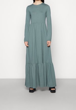 Femme portant une robe longue à manches longues vert sauge avec une taille haute froncée et un ourlet à volants, associée à des sandales à plateforme noires.