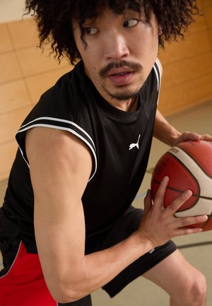 Černý basketbalový dres s bílými akcenty, červené šortky, držící texturovaný basketbal, ukazující atletickou postavu a soustředěný výraz.