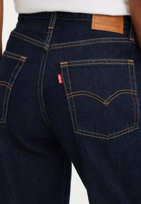 Jeans de mezclilla azul oscuro con costuras amarillas, que presentan dos bolsillos traseros y un parche de cuero con marca en la cinturilla. Etiqueta de logo roja visible.