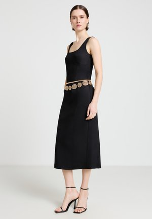 Femme portant une robe midi noire sans manches avec une ceinture décorative en pièces dorées et des sandales noires à talons hauts, debout devant un fond uni.