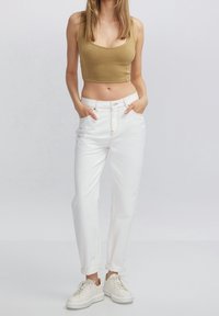 Donna in piedi con le mani in tasca, indossa un top corto color sabbia, jeans bianchi a vita alta e sneakers bianche su uno sfondo neutro.