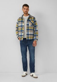 Heren flanellen shirt met een geel, blauw en wit ruitpatroon, kraag, borstzak, gecombineerd met blauwe jeans en witte sneakers.