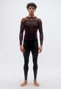 Νεαρός άνδρας στέκεται ξυπόλητος, φοράει μαύρο και πορτοκαλί μακρυμάνικο κορμάκι συμπίεσης και μαύρα κολάν με περίπλοκα σχέδια.