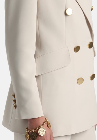 Blazer beige croisé avec de grands boutons dorés, coupe ajustée et tissu à texture lisse. Bracelet chaîne en or avec pendentifs au poignet.