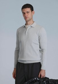 Hellgrau gestreiftes Langarm-Polohemd mit Kragen und drei Knöpfen; kombiniert mit dunklen Hosen und hält eine schwarze Steppjacke.