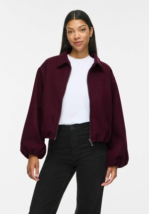 Femme portant une veste zippée bordeaux avec des manches bouffantes, un t-shirt blanc et un pantalon noir taille haute, debout devant un fond uni.