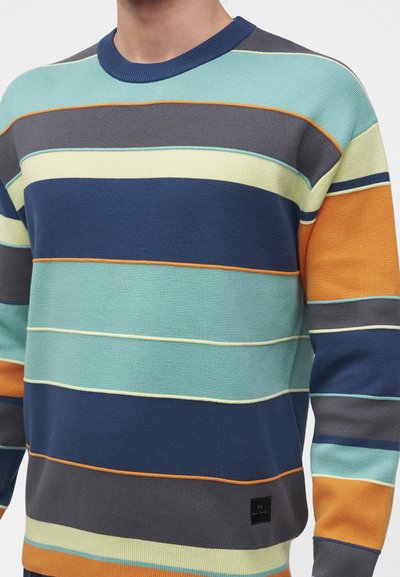 Le haut du corps porte un pull en maille à manches longues avec de larges rayures horizontales bleu, sarcelle, orange, gris et crème.