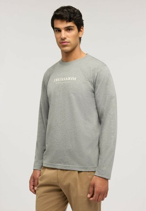 Joven con cabello oscuro que lleva una camisa de manga larga gris claro con el texto "TRUSSARDI GENTLE SOCIETY" y pantalones beige, de pie sobre un fondo liso.