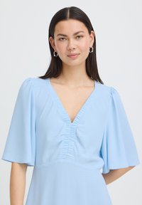 Femme aux cheveux raides et foncés portant des boucles d'oreilles argentées en forme d'anneau et une robe bleu clair à col en V volanté et manches courtes larges.