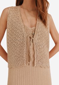 Gilet en tricot beige avec un motif en losange, à l'avant ouvert et aux bords festonnés, superposé sur une robe à côtes assortie. Présente un fermoir décoratif.
