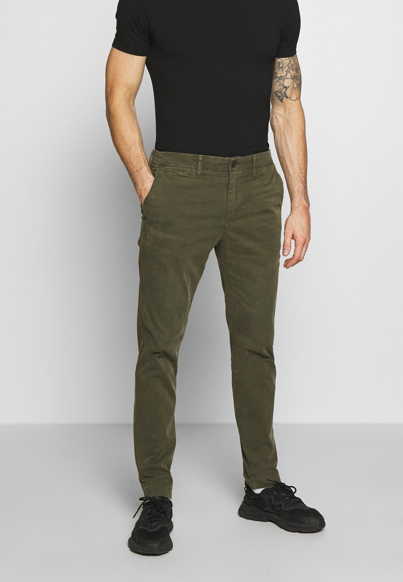 Olive green, slim-fit pants with a button and zipper closure, front pockets, and subtle texture, paired with black sneakers and a fitted black shirt.