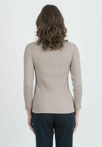 Frau mit lockigem, braunem Haar, die ein beiges, geripptes Langarmoberteil und schwarze Hose trägt, und mit dem Rücken zu einer einfarbigen Wand steht.