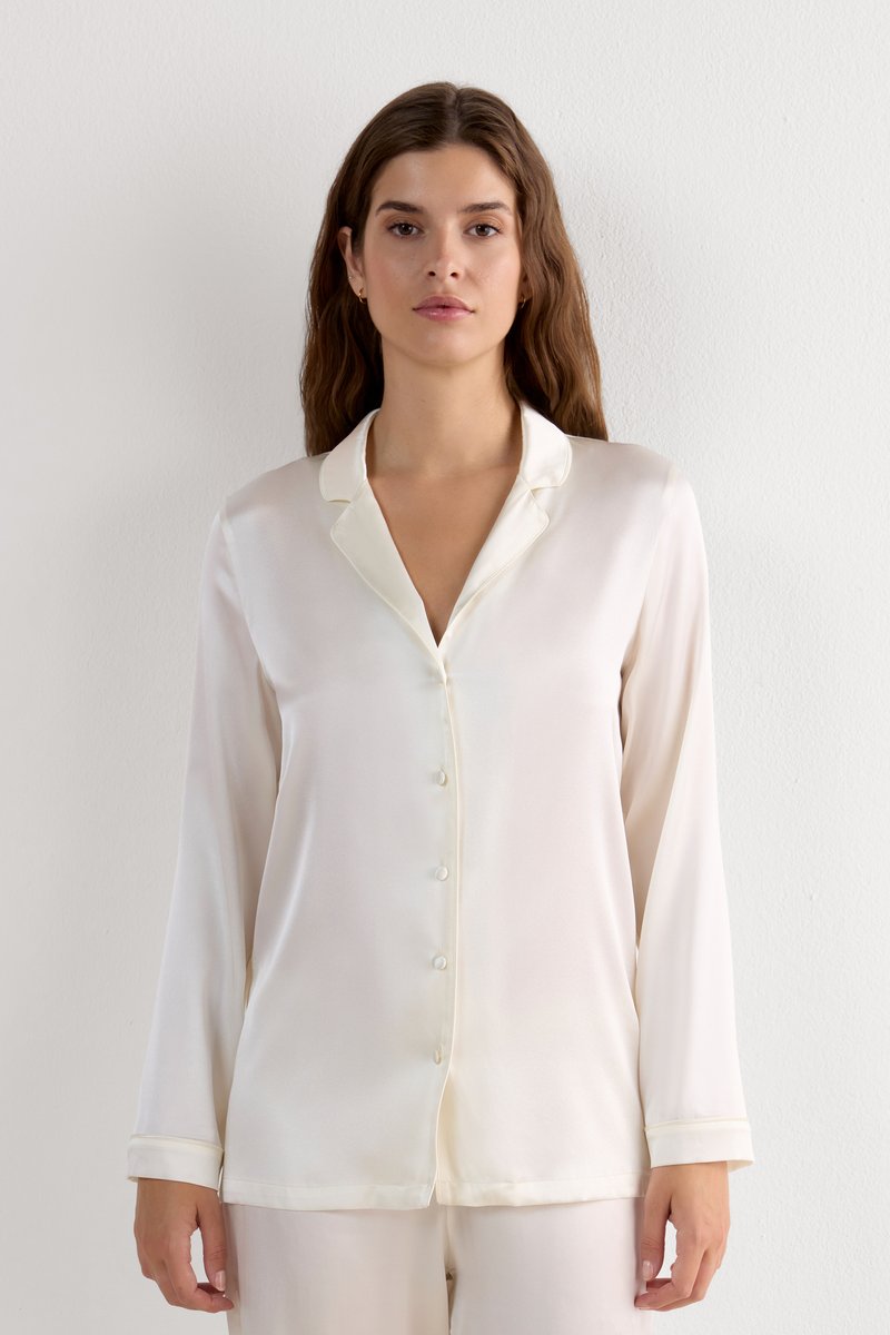 Blusa de seda blanca con cuello, mangas largas y cierre de botones en el frente. Presenta una textura suave y un brillo sutil.
