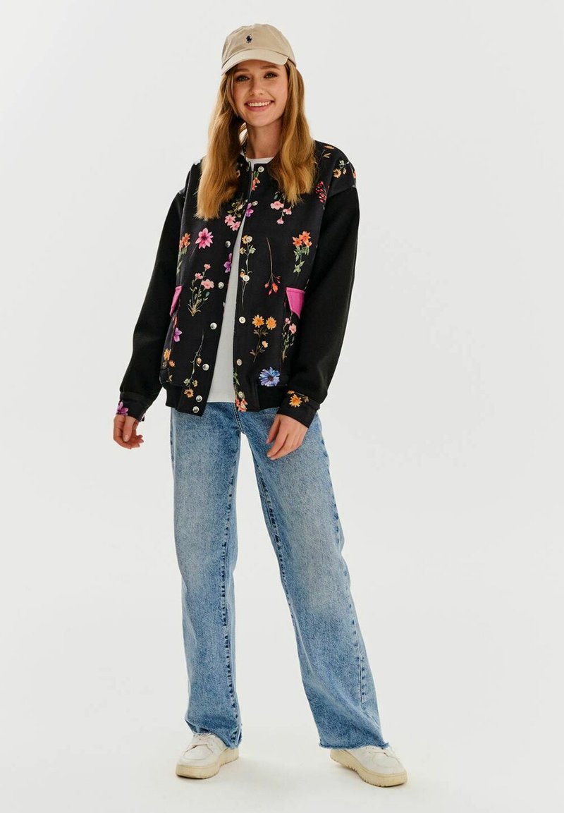 Czarna kurtka bomberka z kolorowym haftem kwiatowym, różowe kieszenie, noszona na białej koszuli i zestawiona z jasnoniebieskimi dżinsami oraz trampkami.