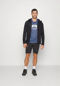 Miesten asu, jossa on tumma teksturoitu takki, sininen T-paita, jossa vuoristokuvio, ja mustat shortsit, yhdistettyinä urheilukenkiin ja valkoisiin sukkiin.