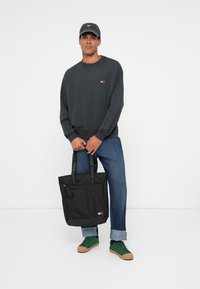 Felpa nera con un piccolo logo, jeans blu, scarpe da ginnastica verdi e una borsa tote nera con spallacci e logo di marca. Sfondo neutro.