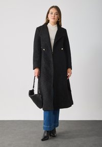 Black textured coat with a long design, double-breasted gold buttons, paired with light-colored top, blue jeans, and black ankle boots.