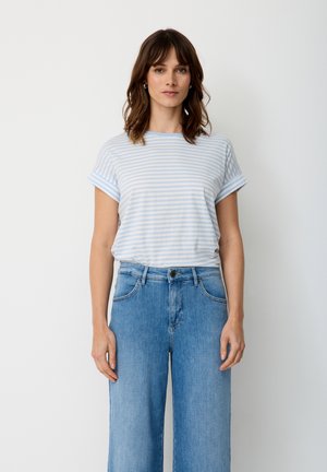 Woman with brown hair wearing light blue and white striped t-shirt and high-waisted blue jeans against plain white background.