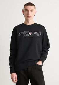 Svart hettegenser med rund hals og lange ermer. Teksten i hvitt og r�ødt sier "GANT 1949 AMERICAN SPORTSWEAR." Laget av bomull.