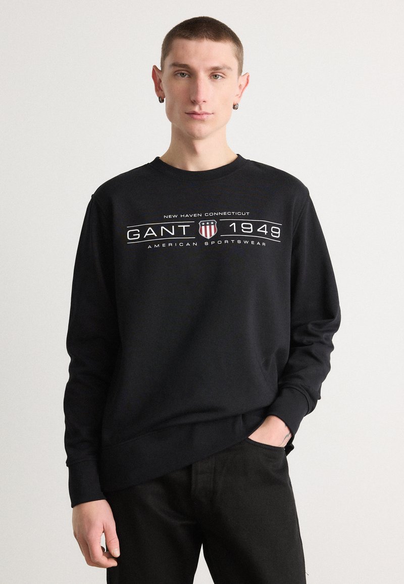 Felpa nera con collo a giro e maniche lunghe. Il testo in bianco e rosso riporta "GANT 1949 AMERICAN SPORTSWEAR." Materiale in cotone.