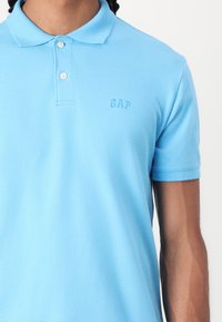 Polo de cor azul clara feito de algodão, com uma pala de três botões e o logo bordado "GAP" no peito. Mangas curtas.