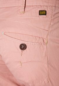 Ljusrosa tygbyxor med en bakficka som har en brun knapp, gula stygn och en G-Star-logotyp. Slät yta, skräddarsydd design.