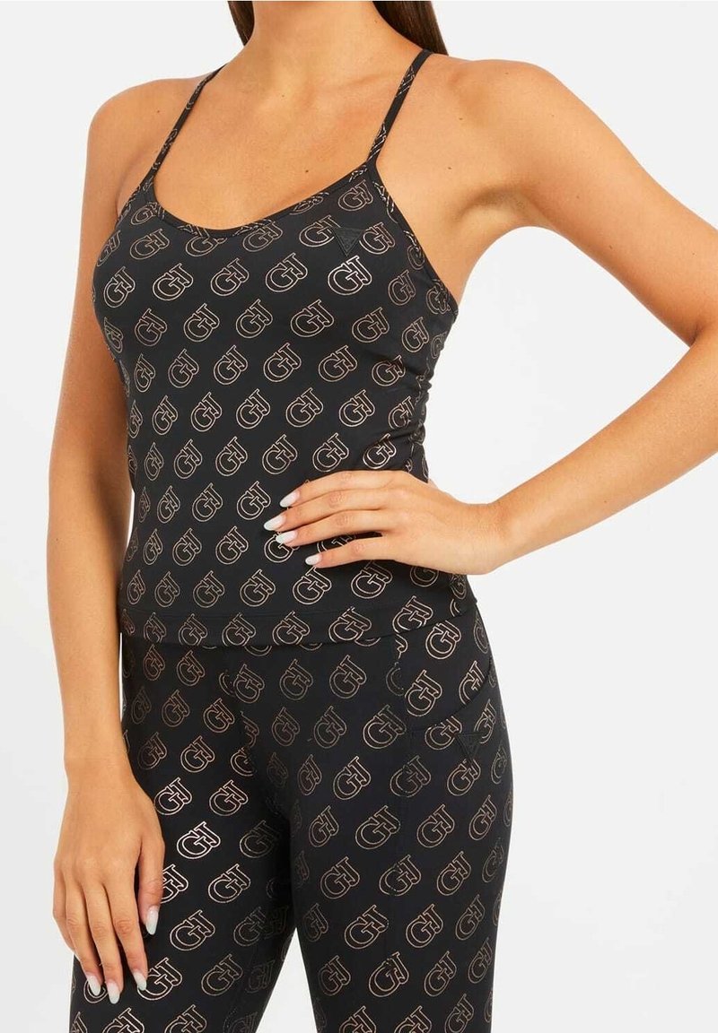 Femme portant un top noir ajusté et un legging avec un motif répétitif de logo, main sur la hanche, sur fond uni.