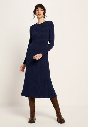 Frau in einem marineblauen, gerippten Strick-Midikleid mit langen Ärmeln und braunen kniehohen Stiefeln, die vor einem einfarbigen Hintergrund steht.