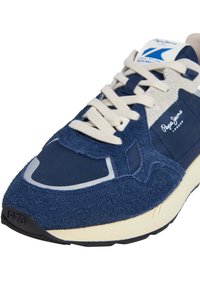 Sapatilha azul marinho com um cabedal texturizado em camurça e nylon, atacadores em creme e uma sola de borracha preta com uma impressão de 1973.
