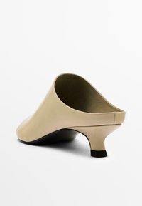 Mules à talons hauts en cuir beige avec un bout rond, un dos ouvert et un accent noir sur le talon. Texture lisse et design minimaliste.