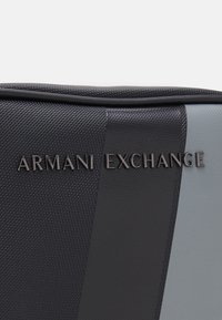 Crna i siva torba s teksturiranom tkaninom, na prednjoj strani s istaknutim logotipom "ARMANI EXCHANGE" u modernom, elegantnom fontu.