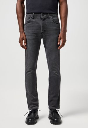 Homme portant un jean noir délavé slim fit, des chaussures noires et une chemise noire, debout devant un arrière-plan uni.