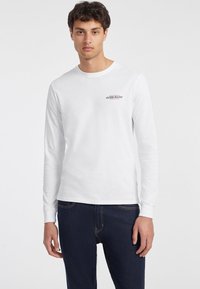 Balta ilgomis rankovėmis marškinėliai, pagaminti iš medvilnės, su mažu raudonu "GUESS JEANS" logotipu viršutinėje kairėje krūtinės pusėje, derinami su tamsiais džinsais.