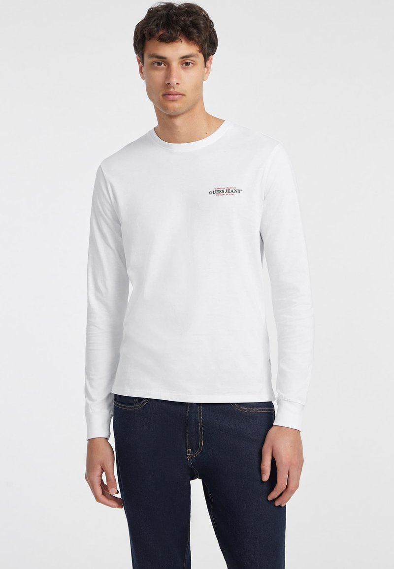 Balta ilgomis rankovėmis marškinėliai, pagaminti iš medvilnės, su mažu raudonu "GUESS JEANS" logotipu viršutinėje kairėje krūtinės pusėje, derinami su tamsiais džinsais.