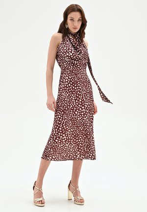 Kobieta w brązowej, bezrękawnikowej sukience midi w białe kropki z wiązaniem przy szyi, w złotych sandałkach na wysokim obcasie, stoi na tle białego tła.