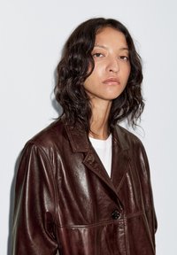 Bruine leren jas met een glanzende afwerking, voorzien van een brede kraag, knoopsluiting en zichtbare stiksel details. Zachte, gestructureerde uitstraling.