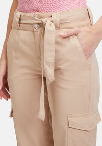 Beige Cargo-Hosen mit gebundenem Bund, zwei Vordertaschen und einer Seitentasche. Glatter Stoff und lockere Passform. Mit zwei Knopfverschluss.