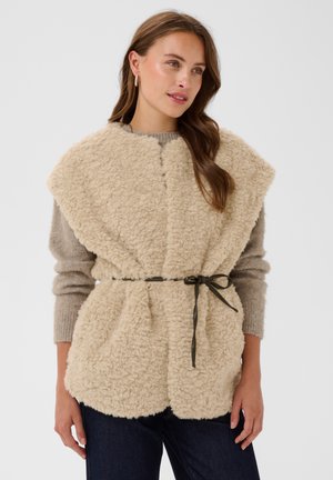 Femme portant un gilet sans manches en fausse peau lainée beige avec une ceinture noire nouée, sur un pull gris et un jean foncé, debout devant un fond neutre.